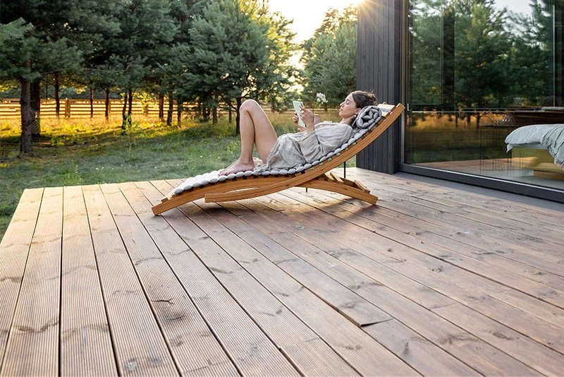 Frau entspannt auf Holzterrasse im Garten, liest bei Sonnenuntergang vor modernem Haus.