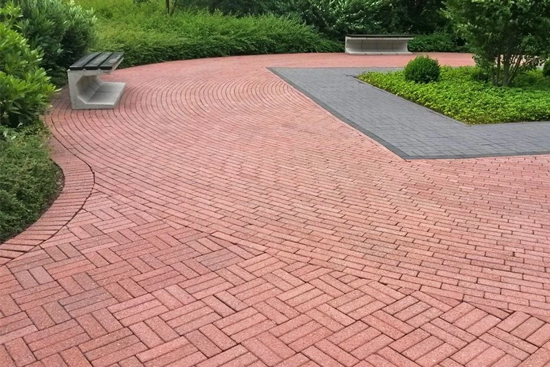 Moderner Gartenweg aus roten Pflastersteinen mit Sitzbänken und grüner Bepflanzung.
